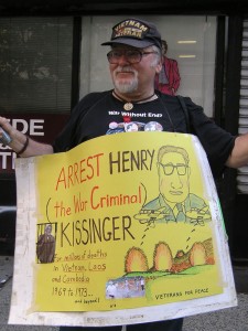 Bill Steyert, wearing a Vietnam Veteran hat and carrying a yellow sign around his neck which says, Arrest Henry (the War Criminal) Kissinger for millions of deaths in Vietnam, Laos, and Cambodia, 1969 to 1973