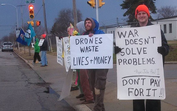 protesters standing along the right side of a road, with signs saying "Drones waste lives and money" and "War doesn't solve problems. - Pope John Paul II ----- Don't pay for it!!!"