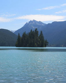 image of an apparent island covered in evergreen trees, with lake water in the foreground and mountains in the background, on a sunny day