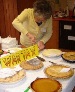 several dessert pies on a table with a sign that says WAR TAX above them. in the background, someone cutting a pie.