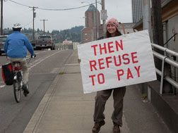 woman on bridge holding sign that reads “then refuse to pay” as bicyclist goes by