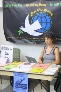 Ari sits at the literature table with a "War No More" dove and earth banner behind her