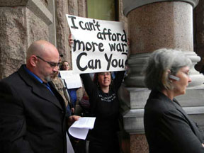 woman holds sign reading “I can’t afford more war. Can you?” as two button-down types walk by