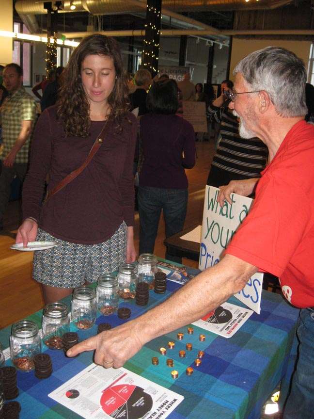 a man behind a penny poll table points at the jars while a woman holding a plate approaches