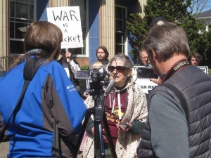 Peg Morton stands in front of a camera talking at a Eugene Tax Day rally in 2015. A sign reading “WAR IS A racket” is held up behind her to the left.