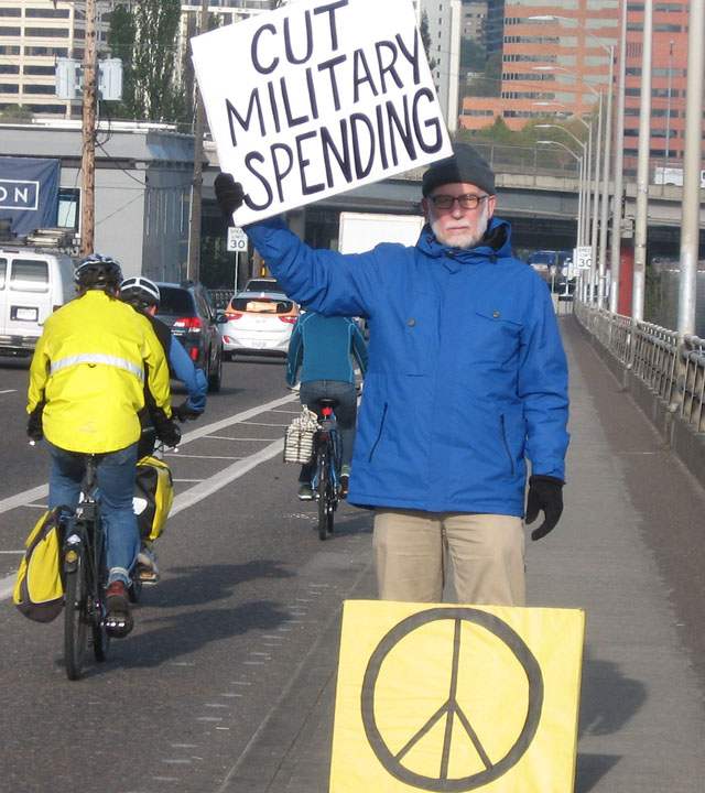 John Grueshow on bridge holding sign reading “Cut Military Spending”