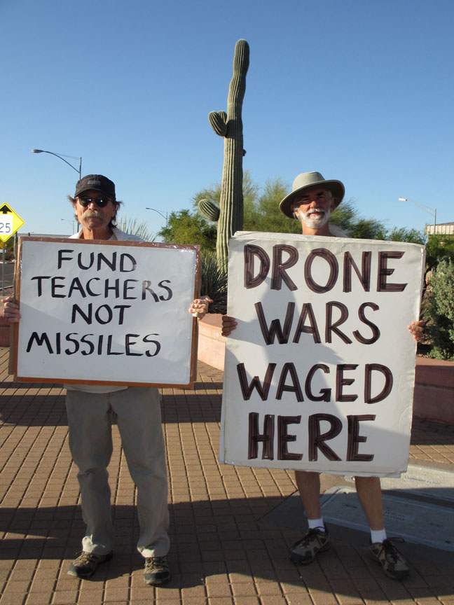 Twi people hold protest signs: “Fund Teachers, not Missile” and “Drone Wars Waged Here”
