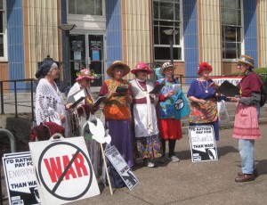 seven members of the Raging Grannies in costume singing outdoors with protest signs nearby