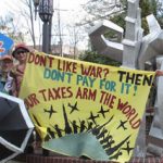 activists holding a black umbrella with a peace sign on it, a blue sign with red letters saying "Honk for peace," and a banner reading "Don't like war? Then don't pay for it! Your taxes arm the world"