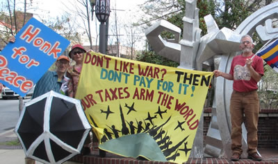 activists holding a black umbrella with a peace sign on it, a blue sign with red letters saying "Honk for peace," and a banner reading "Don't like war? Then don't pay for it! Your taxes arm the world"