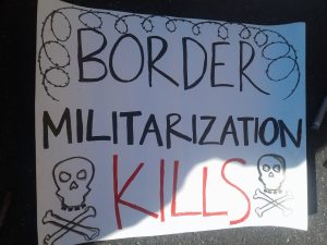 white sign with skull and crossbones and barbed wire, saying in black and red text: "BORDER MILITARIZATION KILLS"