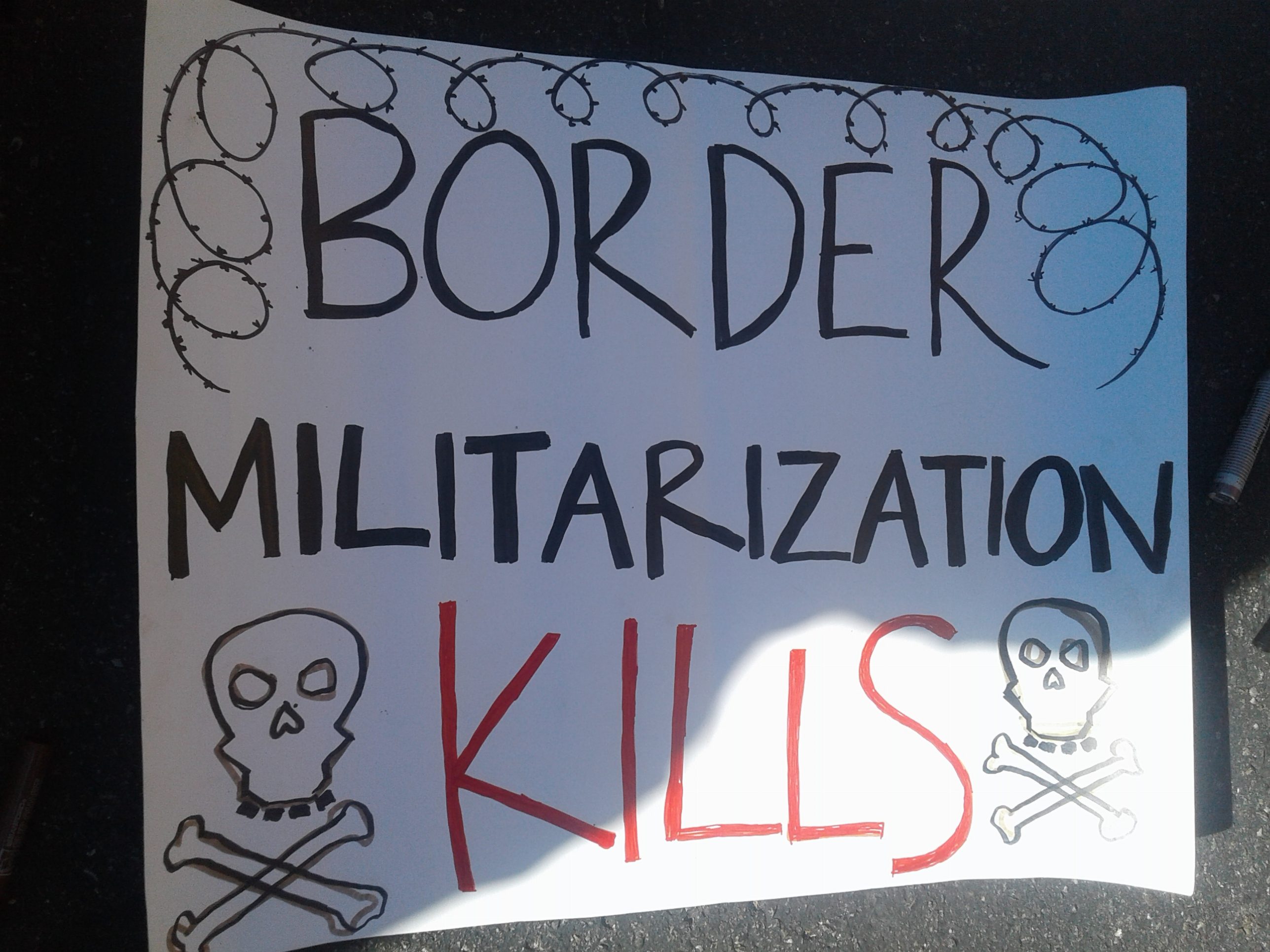 white sign with skull and crossbones and barbed wire, saying in black and red text: "BORDER MILITARIZATION KILLS"