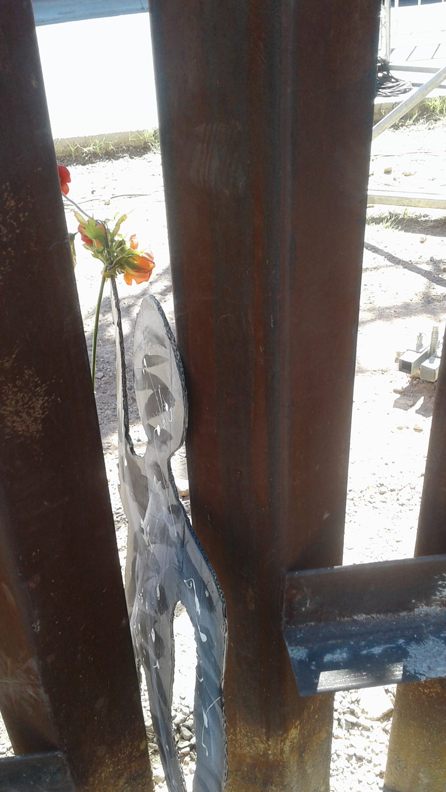 A black cardboard cutout of a person holding a flower slips between the bars of the border wall.