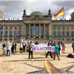 A protest in front of the Reichstag - two people hold a banner reading "BAN URANIUM WEAPONS" in front of a scattered group of about 40 people.