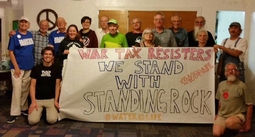 A group of about 15 people standing with a paper banner reading "War Tax Resisters: We Stand with Standing Rock #NoDAPL #waterislife"