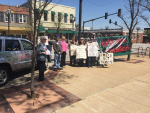 about 15 people holding banners and signs on sidewalk with painted car with messages written on it