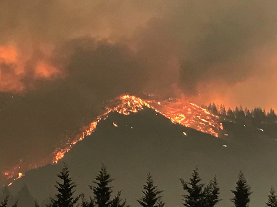 image of mountain tops covered in flames surrounded by smoke and evergreen trees lining the bottom portion of photo