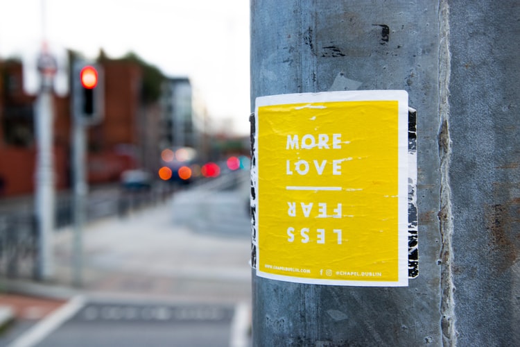 yellow paper posted to a round post with word love more fear less on right hand side and street scene on left side