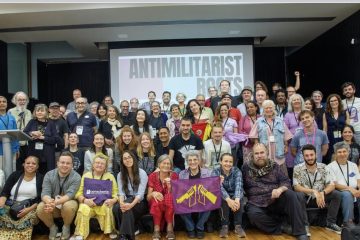 large group of people gathered posing for a photo with screen in background with antimilitarist roots projected